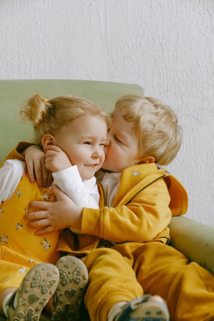 Playful adorable kids in casual outfit sitting together in comfortable armchair and cuddling while having fun in living room